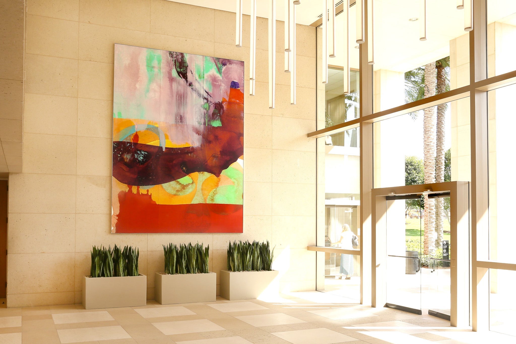 An office interior with a custom abstract painting in shades of red, orange, and green, mounted on a beige wall, with natural light streaming through large glass windows.