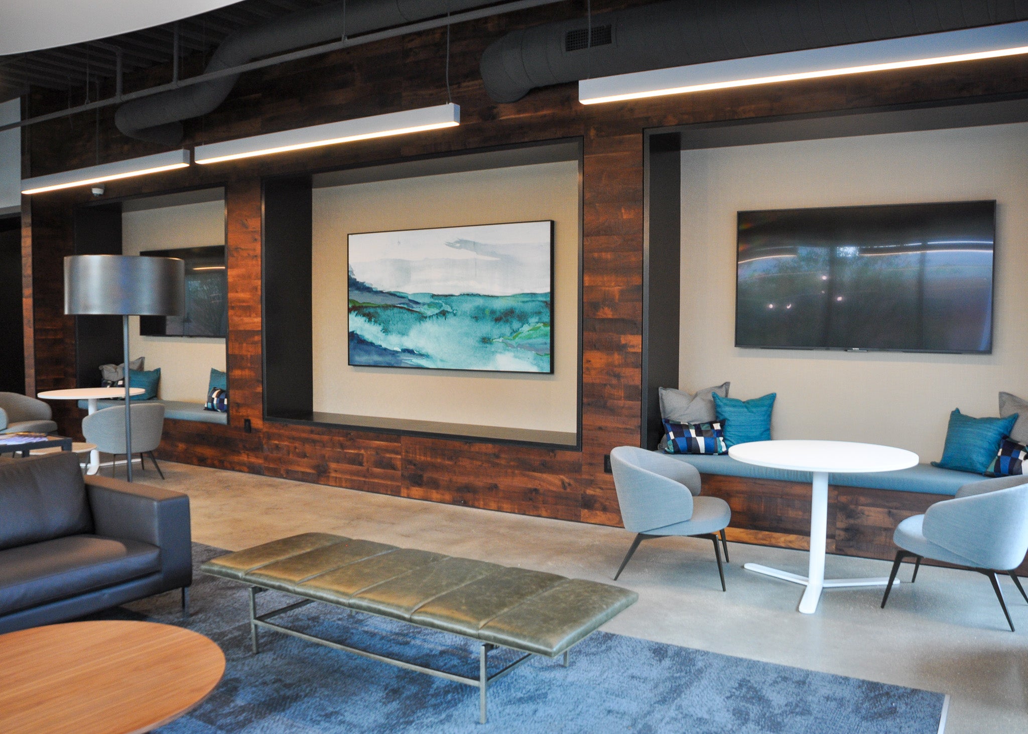 A framed abstract landscape painting by Wrapped Studio, featuring deep blue and teal tones, is mounted in a modern office lounge. The space incorporates wood paneling, contemporary furniture, and cozy seating areas with blue accents that complement the artwork.
