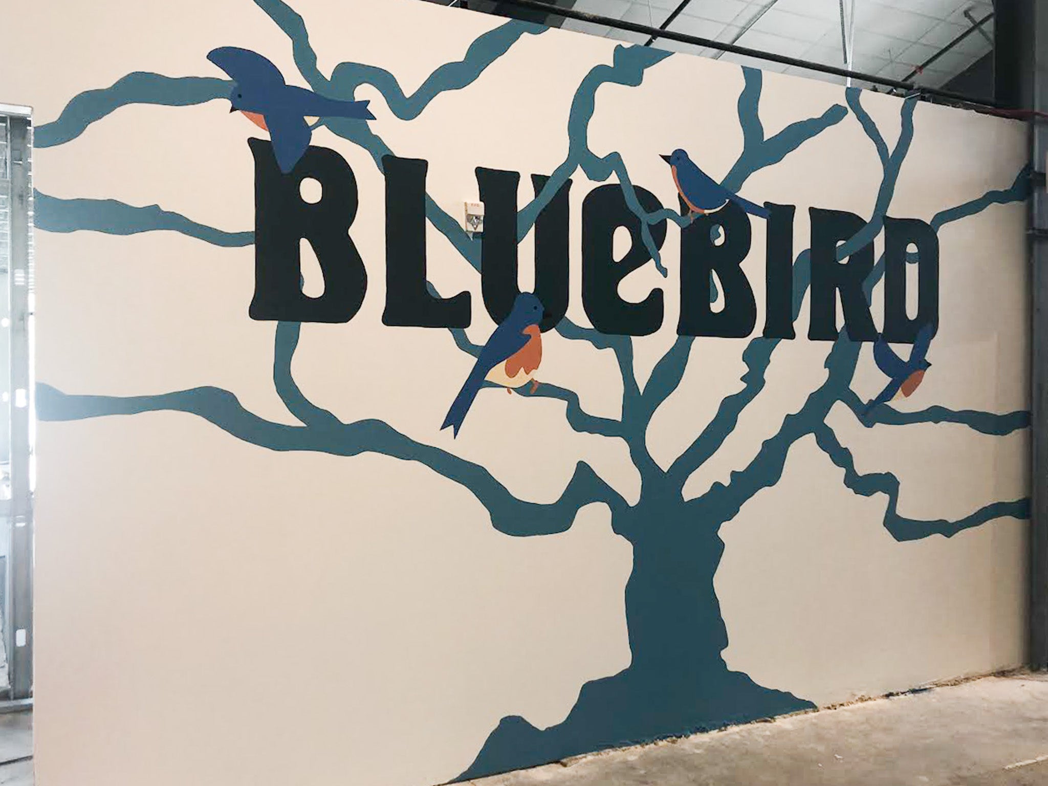 Hand-painted mural by WRAPPED Studio of a branching tree with perched bluebirds and bold lettering, created for Bluebird Market Hall in Silverthorne, CO