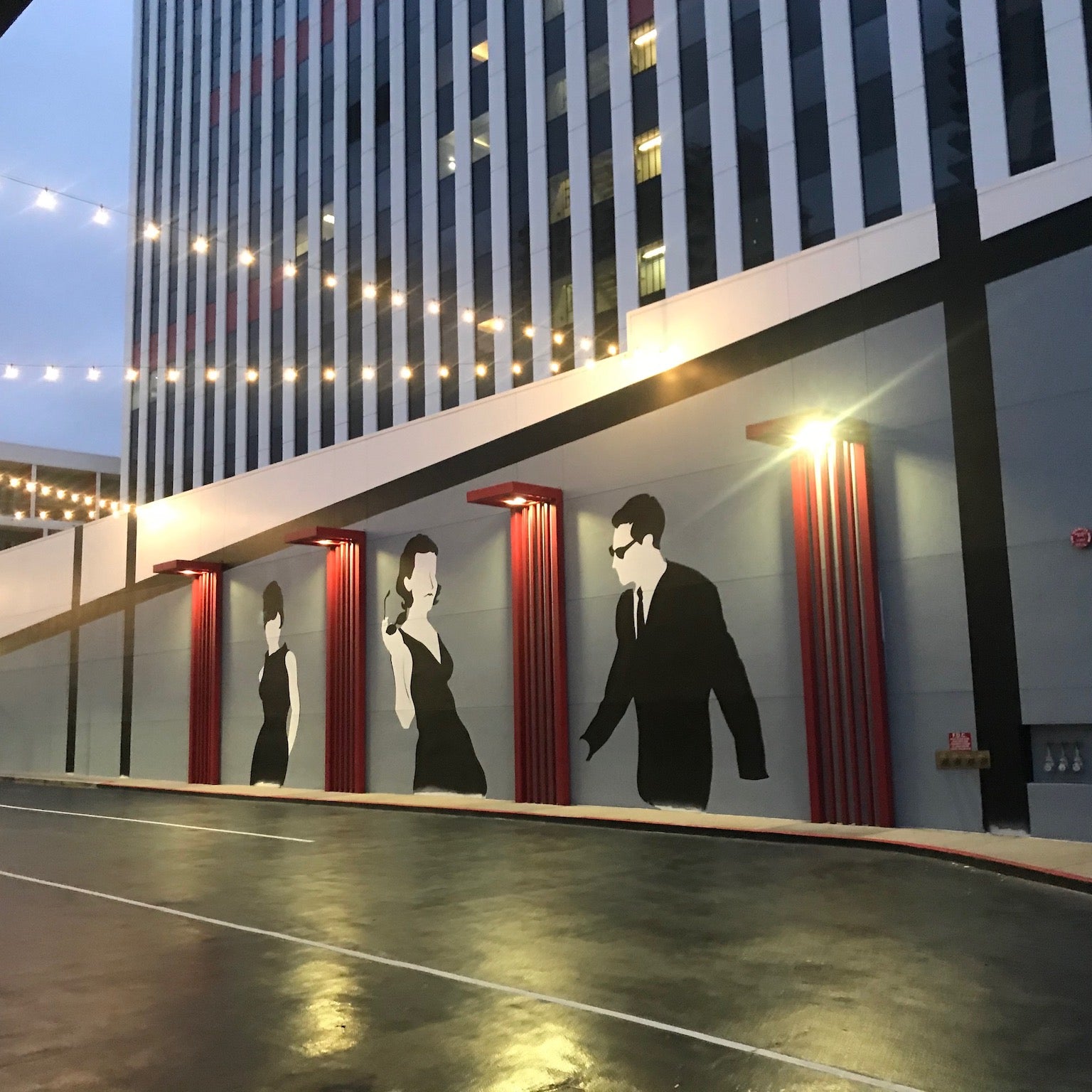 A 100-foot wide hand-painted mural by WRAPPED Studio on the exterior wall of a mid-century modern building, featuring stylized silhouettes of a man and a woman that serve as a local landmark in Long Beach.