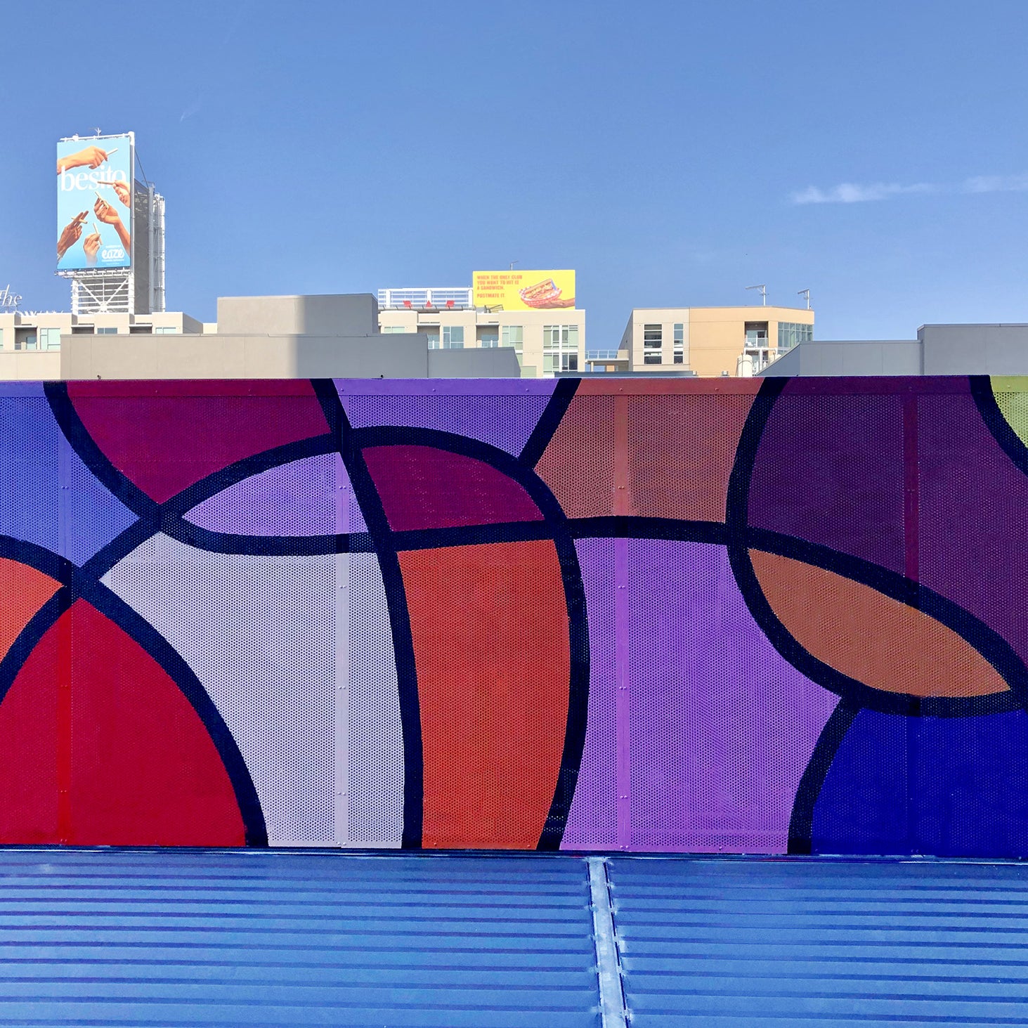 Colorful rooftop mural by WRAPPED Studio on perforated metal, displaying bold abstract patterns in red, purple, and yellow, visible from the surrounding office windows.