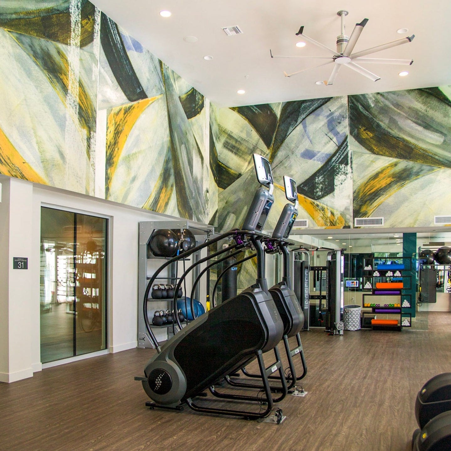A modern gym at Next on Sixth Apartments featuring a custom abstract wall covering with dynamic geometric shapes in green, yellow, and black hues that enhance the contemporary vibe of the fitness area.