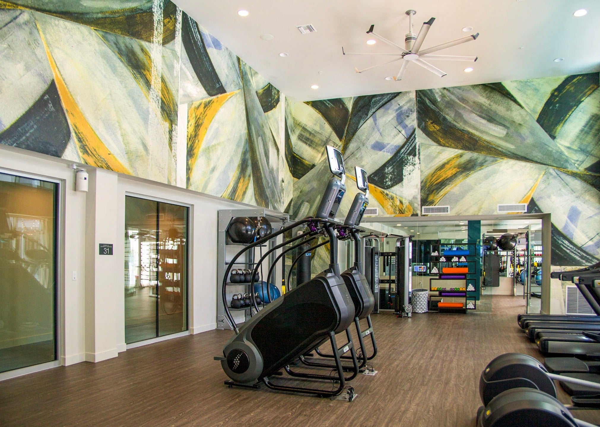 A modern gym at Next on Sixth Apartments featuring a custom abstract wall covering with dynamic geometric shapes in green, yellow, and black hues that enhance the contemporary vibe of the fitness area.
