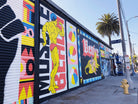 Colorful street mural by WRAPPED Studio on the exterior of Charles Arnoldi's art studio in Venice, featuring a dynamic collage of local icons, including a depiction of Arnold Schwarzenegger.