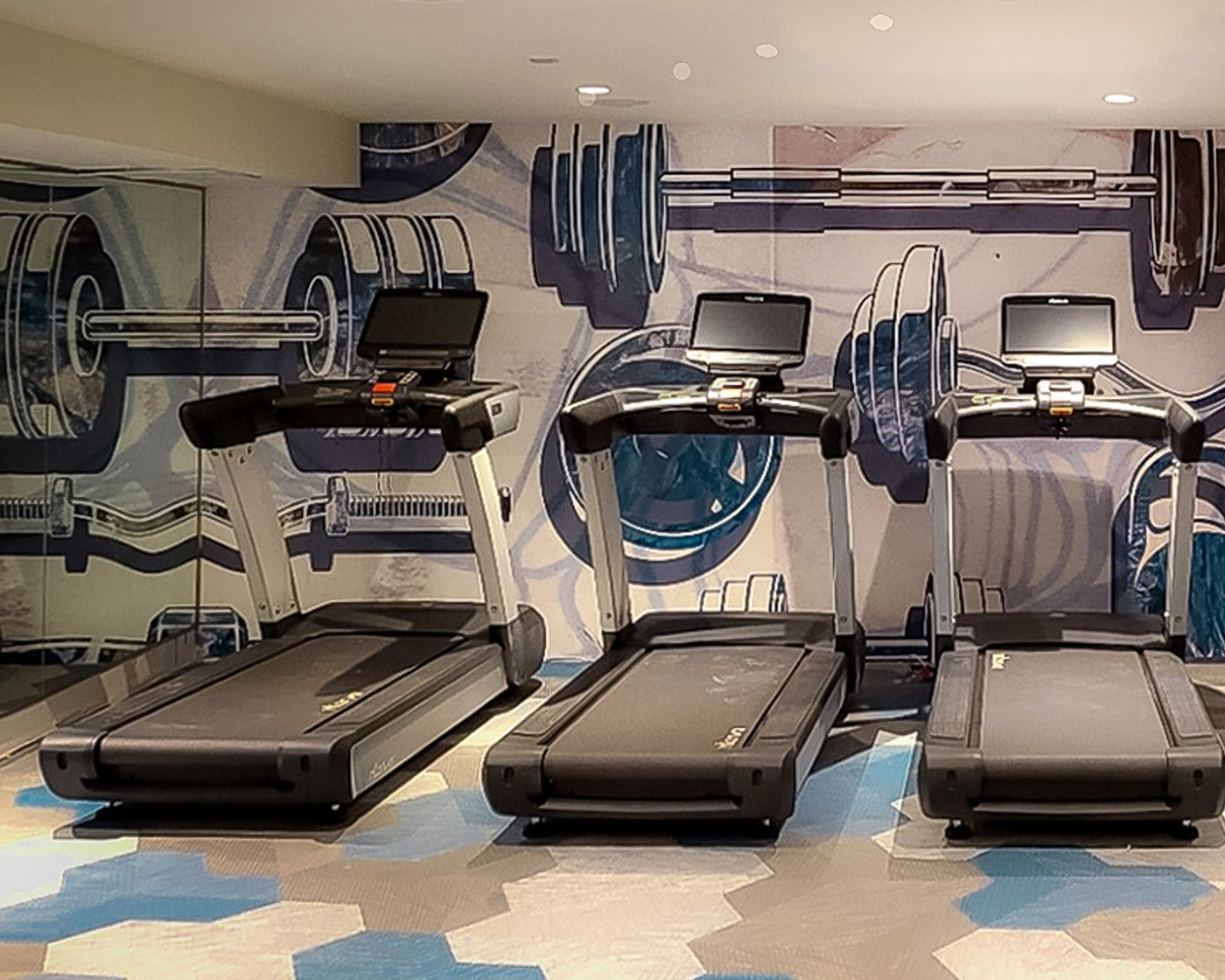 Custom fitness center mural in a multifamily building, featuring oversized illustrations of barbells and dumbbells in blue and gray tones. Positioned behind a row of treadmills, the mural enhances the gym’s dynamic and modern aesthetic.