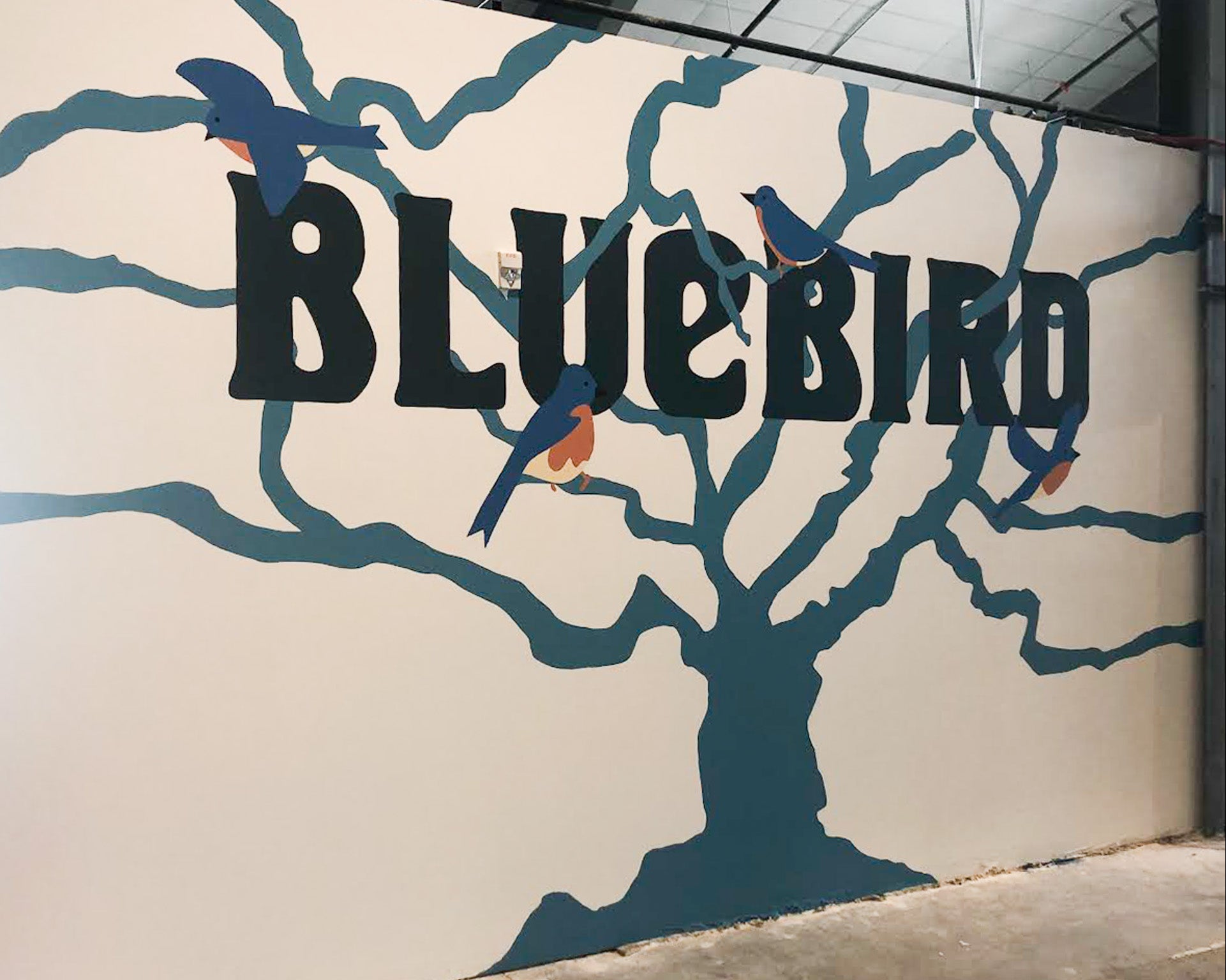 Hand-painted mural by WRAPPED Studio of a branching tree with perched bluebirds and bold lettering, created for Bluebird Market Hall in Silverthorne, CO