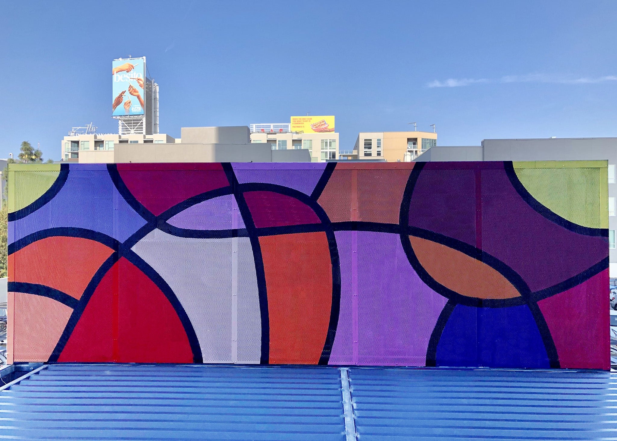 Colorful rooftop mural by WRAPPED Studio on perforated metal, displaying bold abstract patterns in red, purple, and yellow, visible from the surrounding office windows.